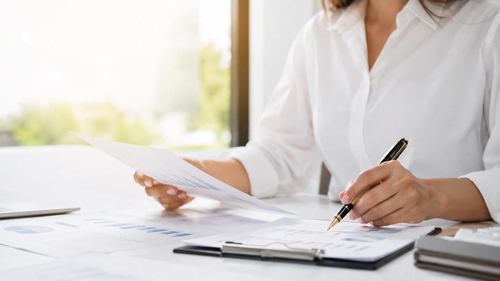 Professional bookkeeper reviewing financial reports and writing notes at a bright office desk.